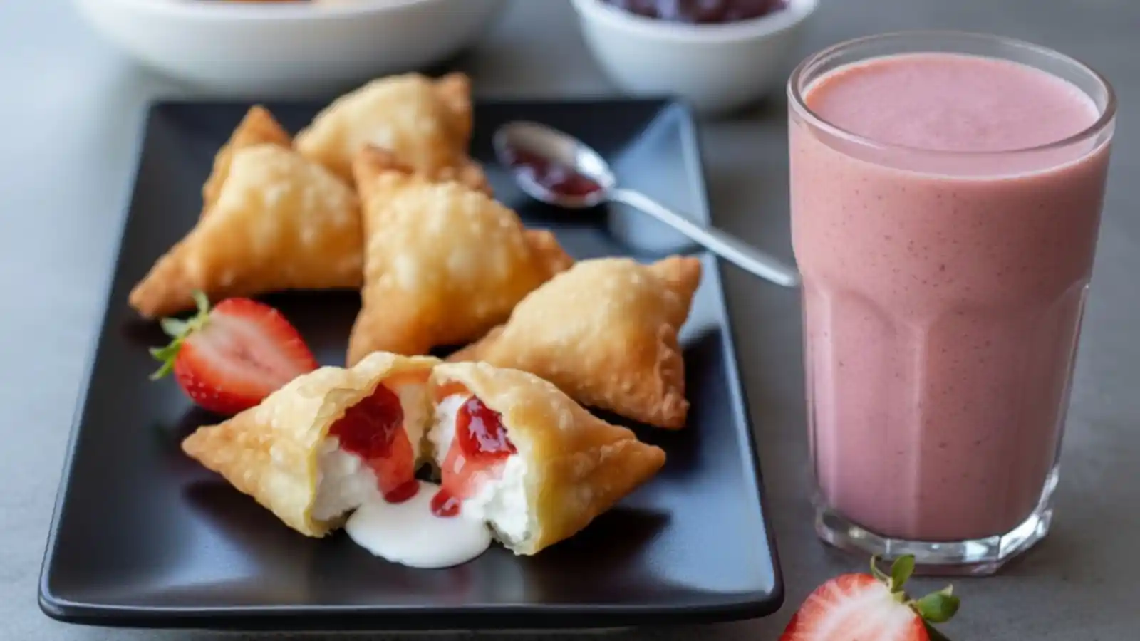 Golden-brown deep fried strawberry cheesecake stuffed wonton bites on a black plate, with one sliced open to show the creamy cheesecake and strawberry filling and a strawberry smoothie aside