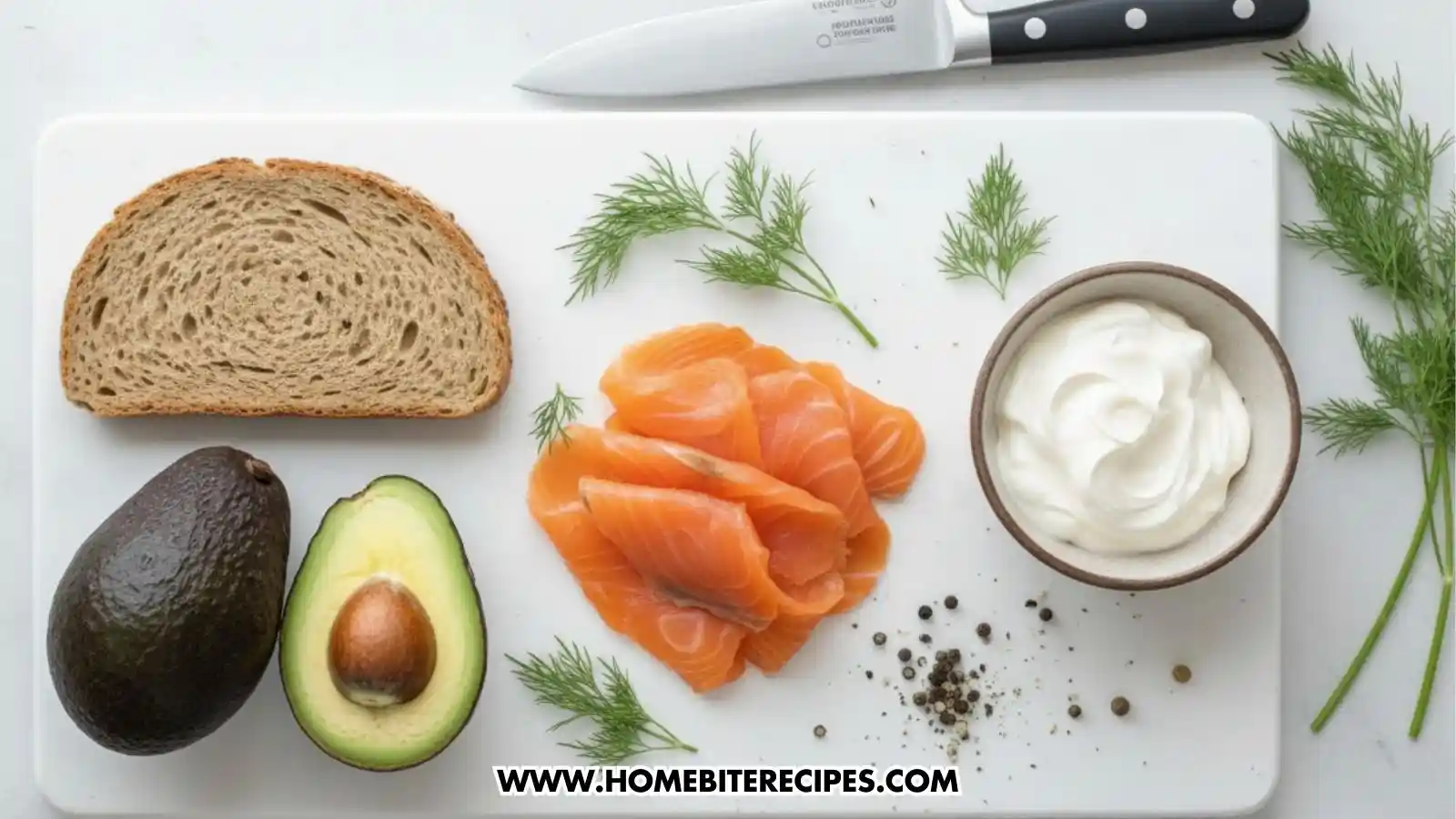 A collection of fresh ingredients for smoked salmon avocado toast arranged on a white cutting board. The ingredients include a slice of rustic whole-grain bread, a whole avocado next to a halved avocado with the pit visible, several ribbons of bright pink smoked salmon, a small bowl of thick white cream sauce, fresh dill sprigs, and scattered black peppercorns. A chef's knife rests at the top of the frame.