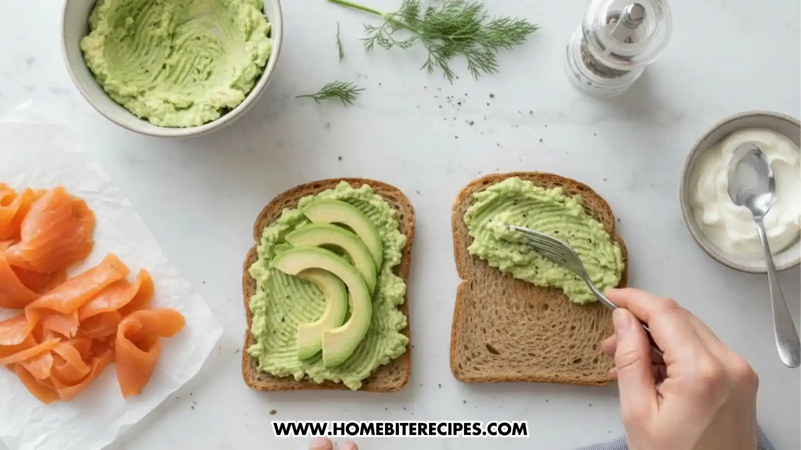 A top-down view of the preparation process for smoked salmon avocado toast. One slice of toasted grain bread is partially spread with mashed green avocado using a fork, while a second slice next to it is fully covered and topped with three fresh avocado slices. Surrounding the toast are bowls of extra mashed avocado, creamy white sauce, a pile of smoked salmon, fresh dill sprigs, and a pepper grinder.