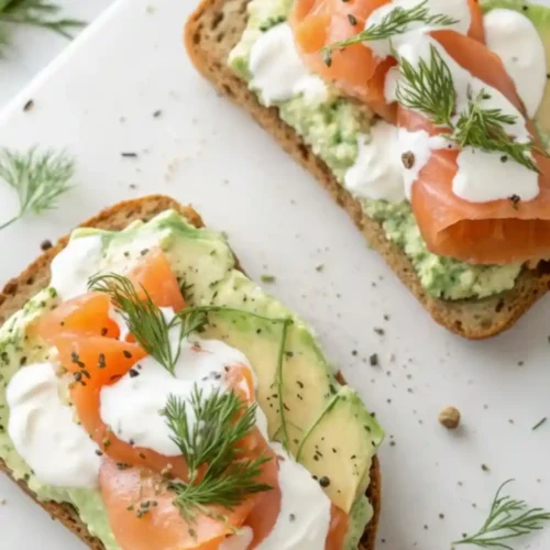 Overhead view of two slices of avocado toast topped with smoked salmon, dollops of creamy dill sauce, and fresh herbs on a white board.