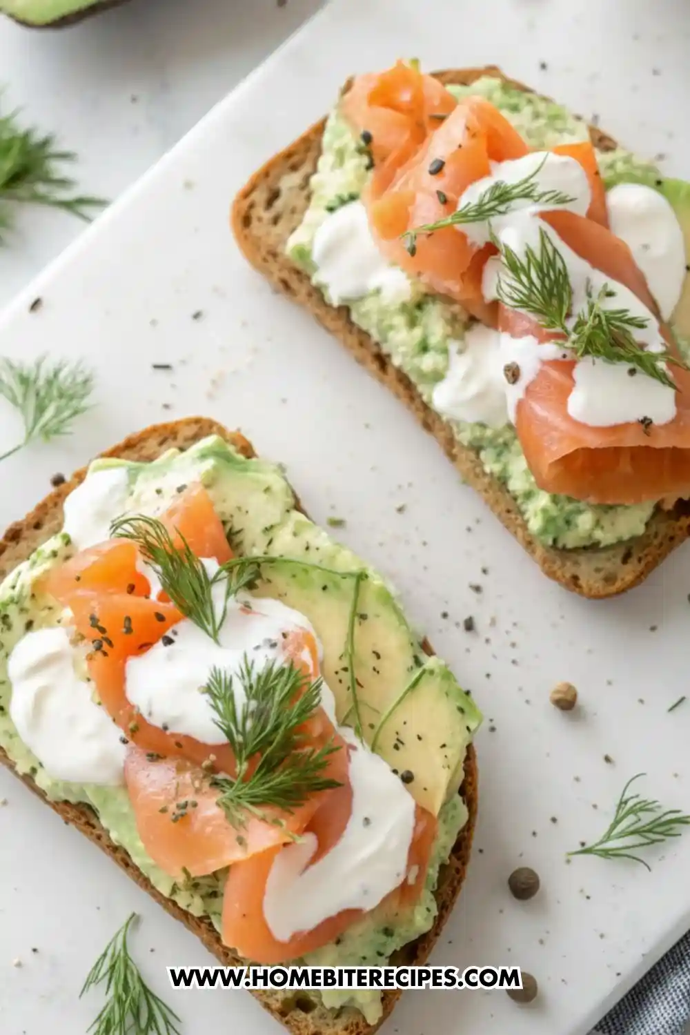 Overhead view of two slices of avocado toast topped with smoked salmon, dollops of creamy dill sauce, and fresh herbs on a white board.