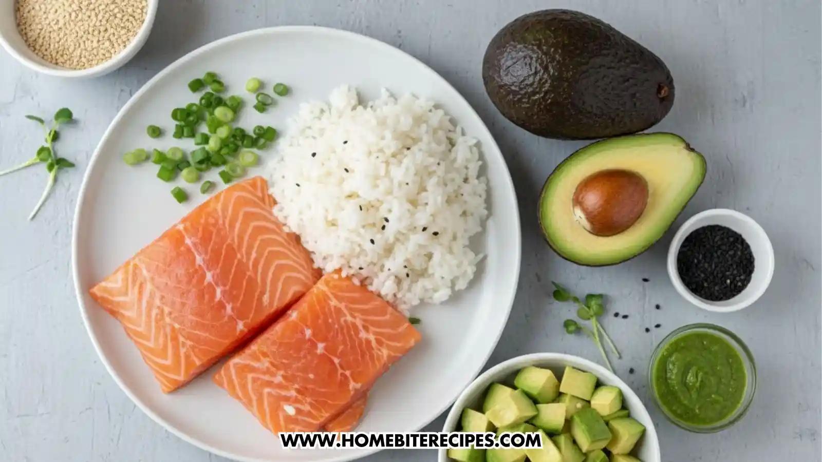 A top-down view of fresh ingredients for a sushi tower arranged on a grey surface. A white plate holds two raw salmon fillets, a mound of white sushi rice seasoned with black sesame seeds, and a pile of chopped green onions. Surrounding the plate are a whole avocado, an avocado half with the pit, small bowls of white and black sesame seeds, a bowl of diced avocado, and a small container of green wasabi or avocado sauce.