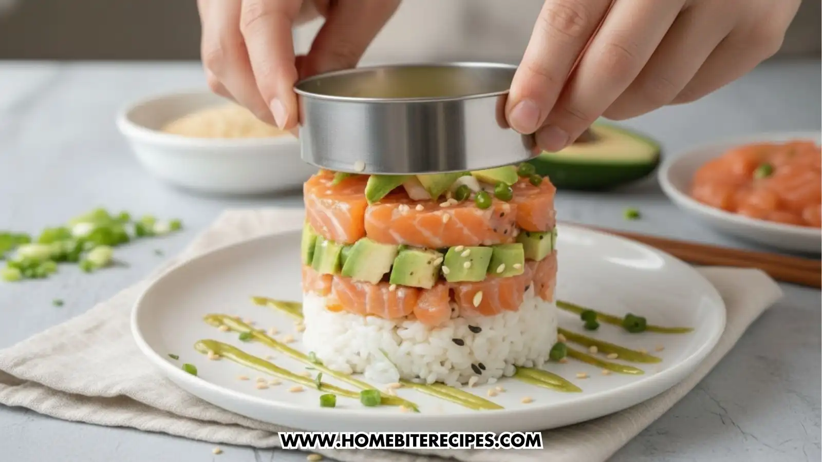 A close-up shot of a salmon and avocado sushi tower being assembled using a circular metal mold. The base layer is white sushi rice, followed by a layer of cubed salmon, then a layer of diced avocado, and a final top layer of salmon. A person's hands are lifting the metal ring to reveal the perfectly stacked layers. The plate is decorated with diagonal stripes of green sauce and a sprinkle of sesame seeds.