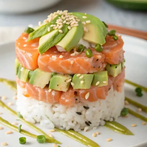 A gourmet stacked sushi tower on a white plate with layers of white sushi rice, diced fresh salmon, and cubed avocado, topped with avocado slices and sesame seeds.