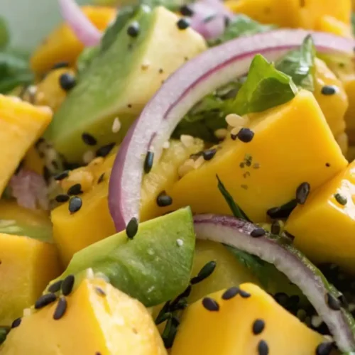 Fresh mango avocado salad with ripe mango cubes, avocado chunks, mixed greens, red onion, sesame seeds, and chili flakes in a ceramic bowl.
