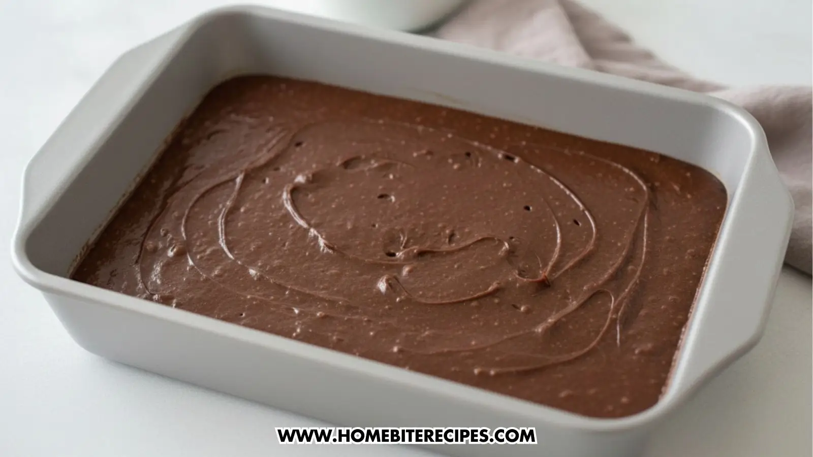 Smooth, dark chocolate brownie batter spread evenly in a rectangular grey baking pan. The batter has a glossy finish with slight swirl marks on the surface before being baked.