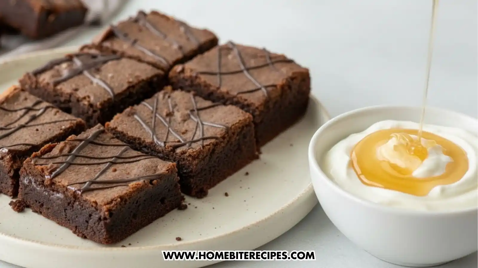 Six square, flourless Greek yogurt brownies arranged on a ceramic plate, drizzled with dark chocolate. To the right, a small white bowl contains thick Greek yogurt topped with a swirl of honey being poured from above.